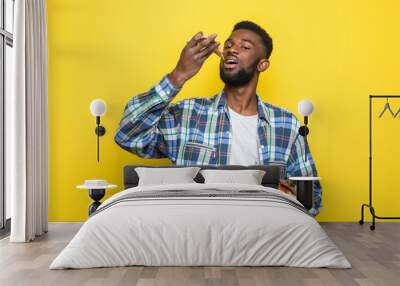 African american man eating slice of pizza standing over isolated yellow background Wall mural